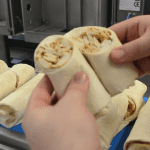 Close-up of hands holding a wrap sliced at a 45-degree angle, showing clean ultrasonic cuts, with more wraps on a conveyor in the background.