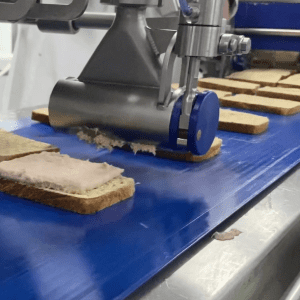 Close-up of a Volumetric Sauce Depositor (VSD) sheeting a tuna mix evenly onto a slice of bread.