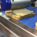 Close-up of a Volumetric Sauce Depositor (VSD) sheeting a tuna mix evenly onto a slice of bread.