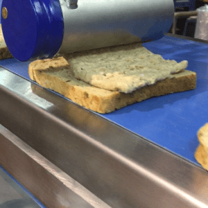 Close-up of a Volumetric Sauce Depositor (VSD) sheeting a tuna mix evenly onto a slice of bread.