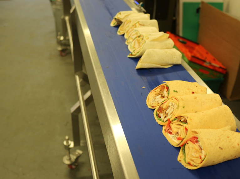 Freshly sliced wraps arranged in rows on a blue conveyor belt, showing clean, even cuts through various filled tortillas.
