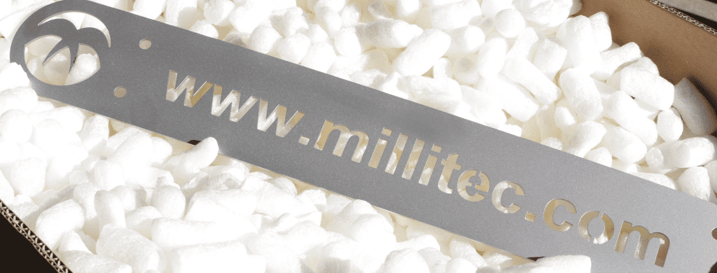 Close-up of a precision laser-cut metal sign featuring the Millitec logo and website address, displayed on a bed of white packing peanuts. Showcasing high-quality metal fabrication for branding and industrial equipment.